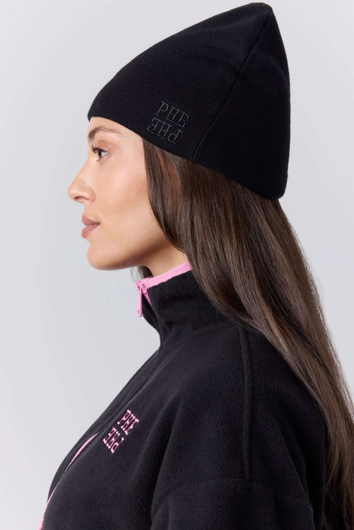 A woman in profile wearing a black knit beanie with "PHE PHE" embroidered on it. She is wearing a black fleece jacket with pink piping. Photographed against a gray studio wall.