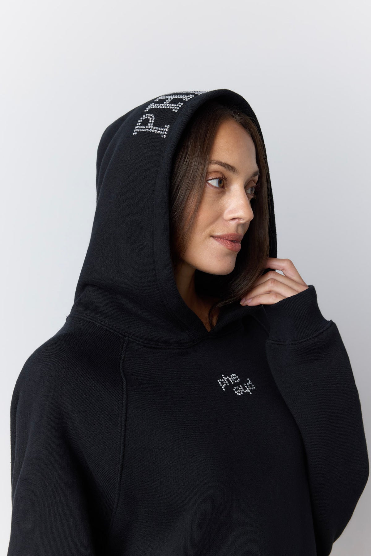 A woman wearing a black pullover hoodie with silver rhinestone lettering on the hood and chest, photographed against a light gray studio background.