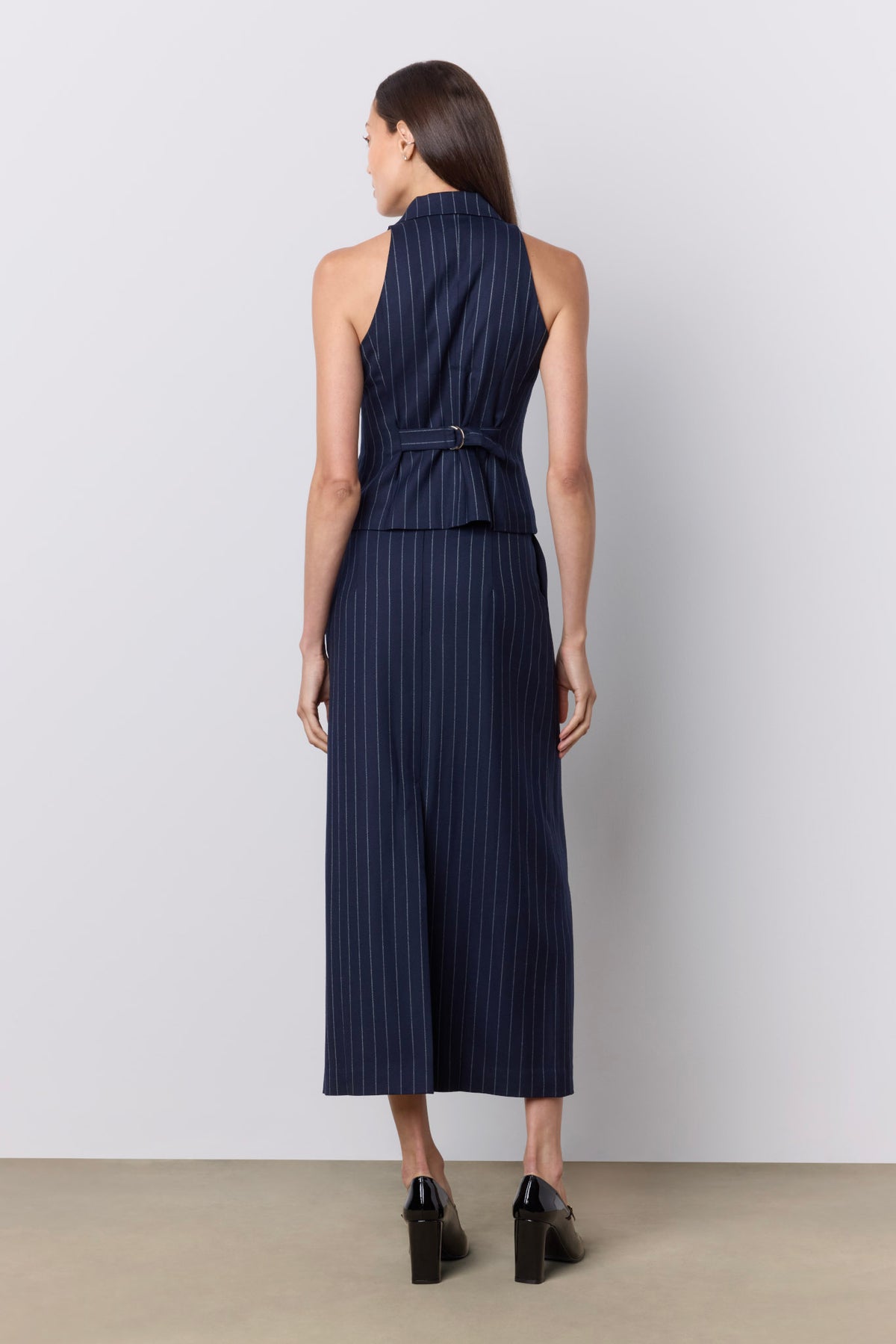 A woman viewed from the back wearing a sleeveless navy vest with thin vertical white pinstripes, a high neckline, and a matching fabric tie belt at the waist. She is standing against a plain light gray studio wall on a beige floor.