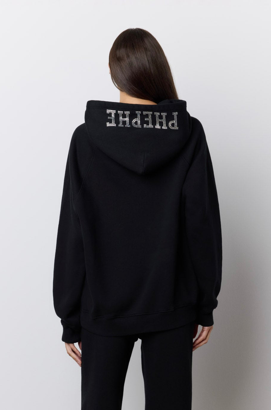 A model viewed from behind wearing a black hoodie and matching wide-leg pants with rhinestone 'PHEPHE' lettering on the hood and legs, standing against a plain light gray wall.