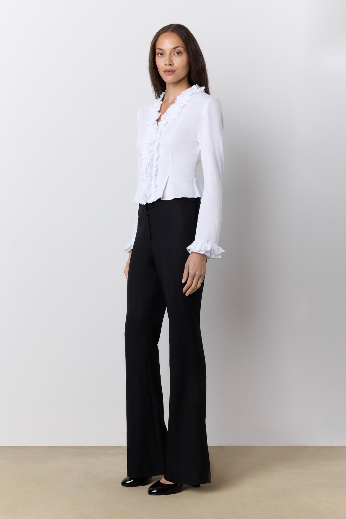 A woman modeling a white long-sleeve blouse with ruffled trim and black wide-leg trousers against a plain light gray studio background.