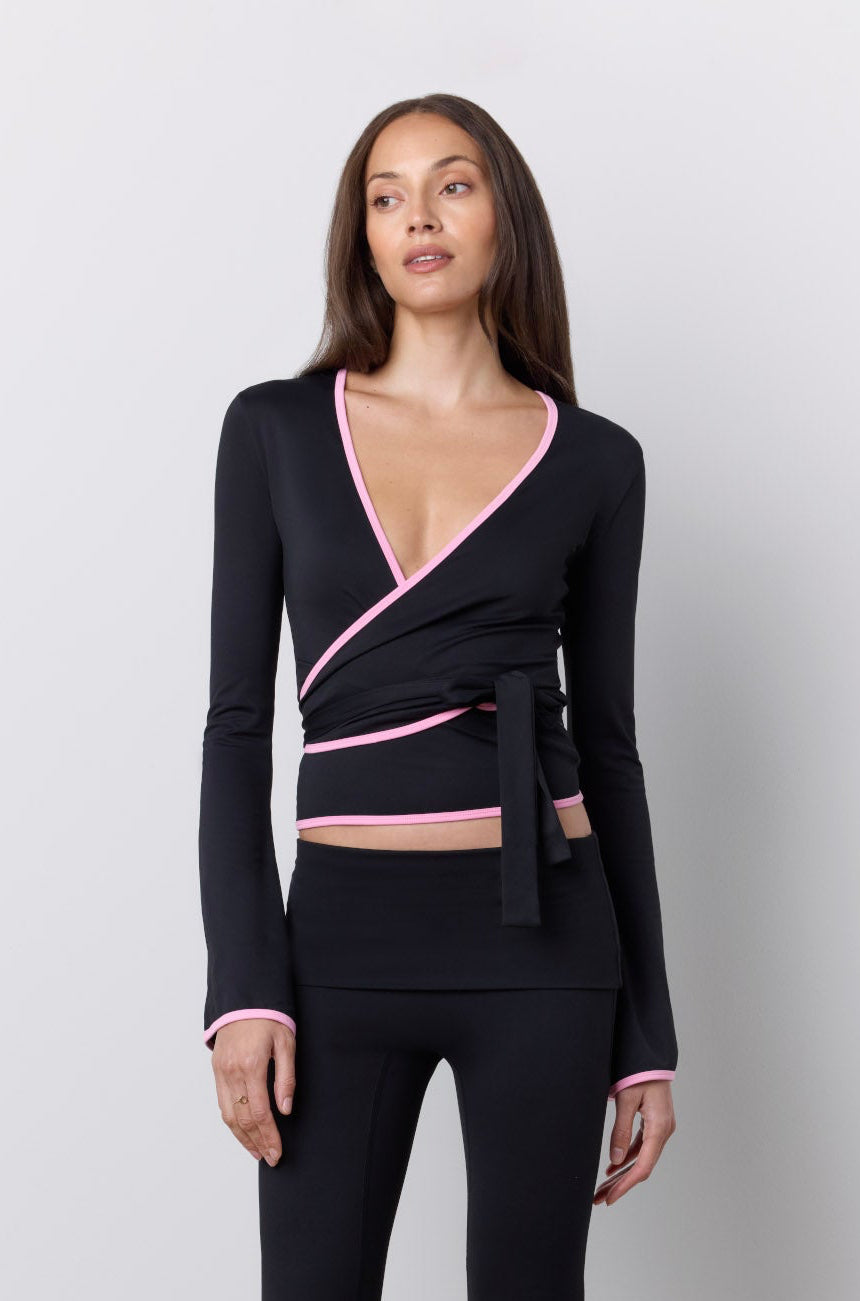 A woman modeling a black wrap-style long-sleeve top and matching flared pants set with pink trim details, standing against a light gray studio background.