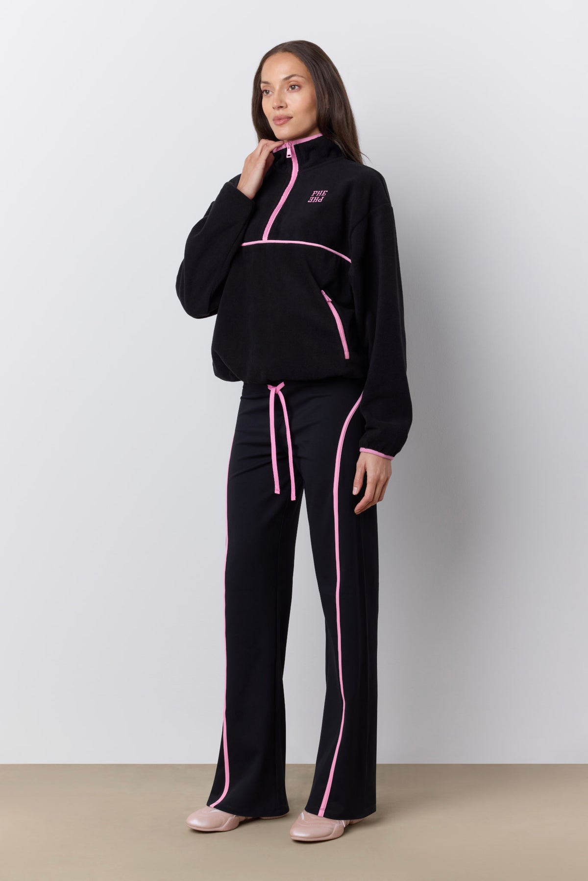 A woman modeling a black tracksuit with bright pink piping and a pink drawstring, standing against a light gray studio wall.