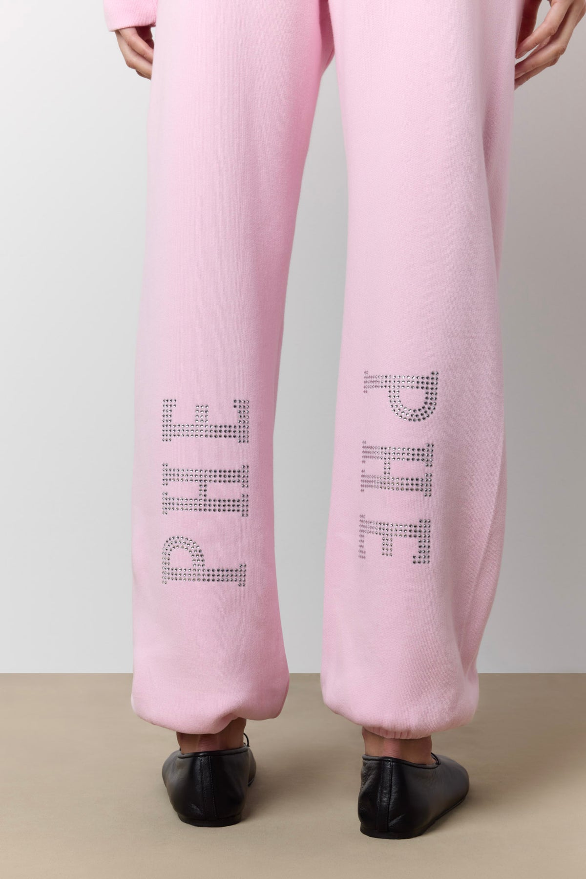 Pink sweatpants with 'PHE' lettering in a rhinestone-like pattern on each leg, shown from the back on a model wearing black shoes.