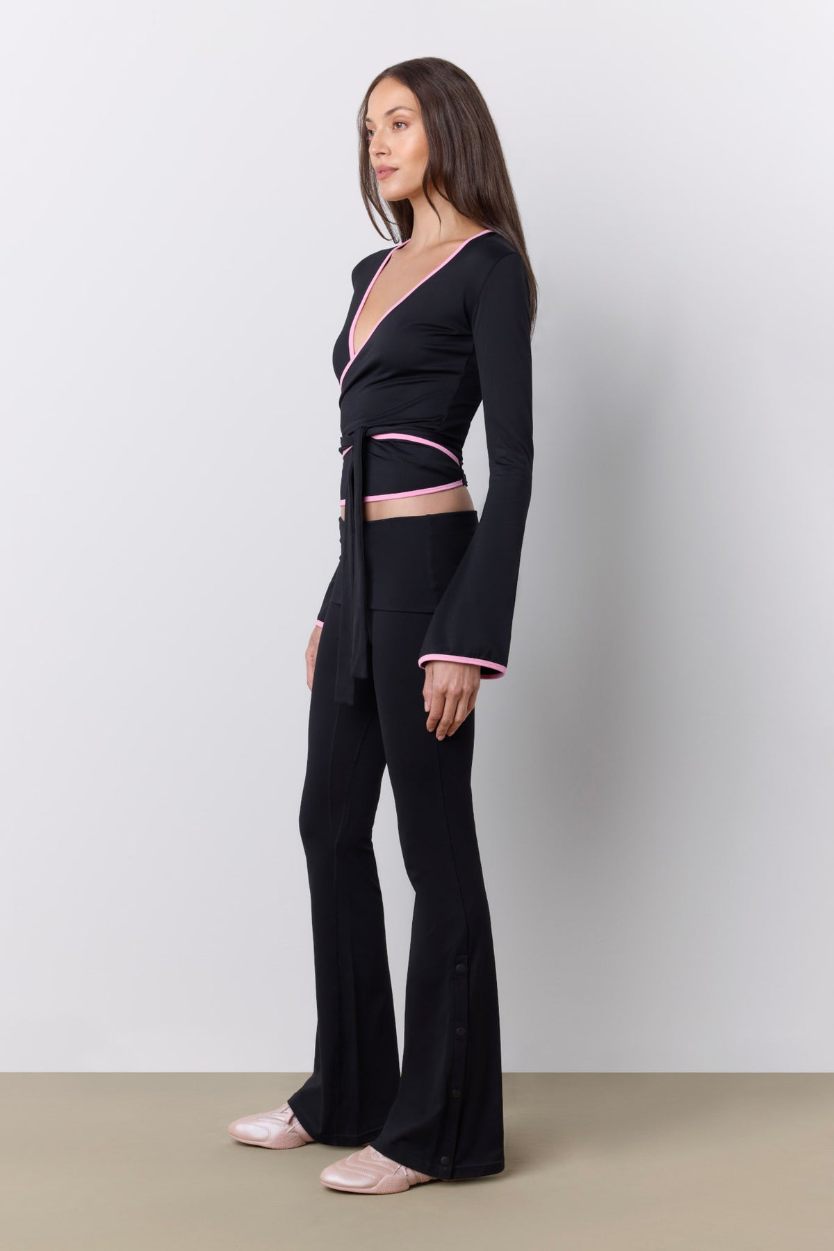 A woman modeling a black long-sleeve wrap-style top and matching flared pants set with thin pink trim, standing against a light gray studio wall.
