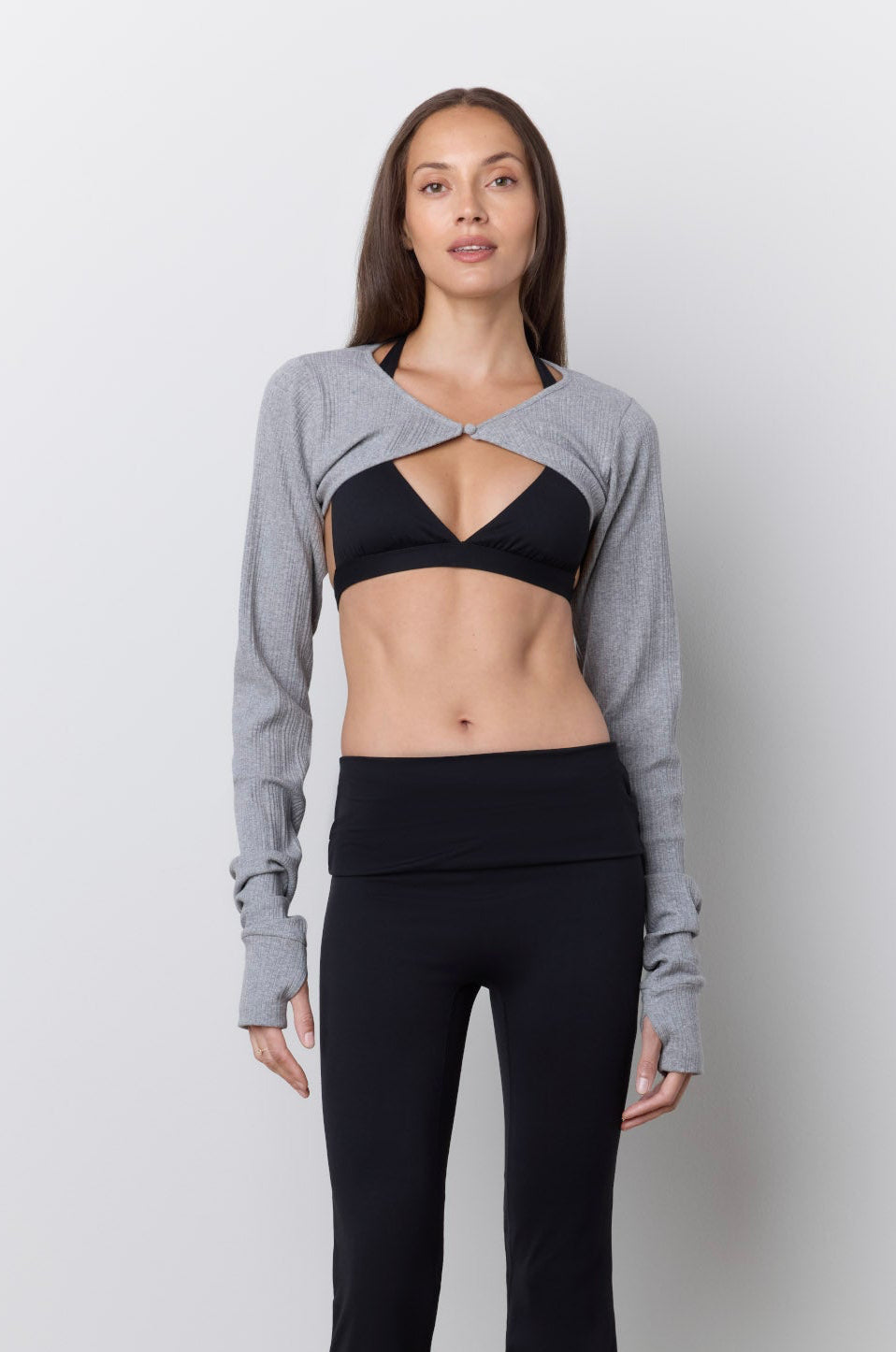 A woman stands against a light gray wall wearing a gray long-sleeve bolero style top over a black sports bra, paired with black flared leggings and pink sneakers.