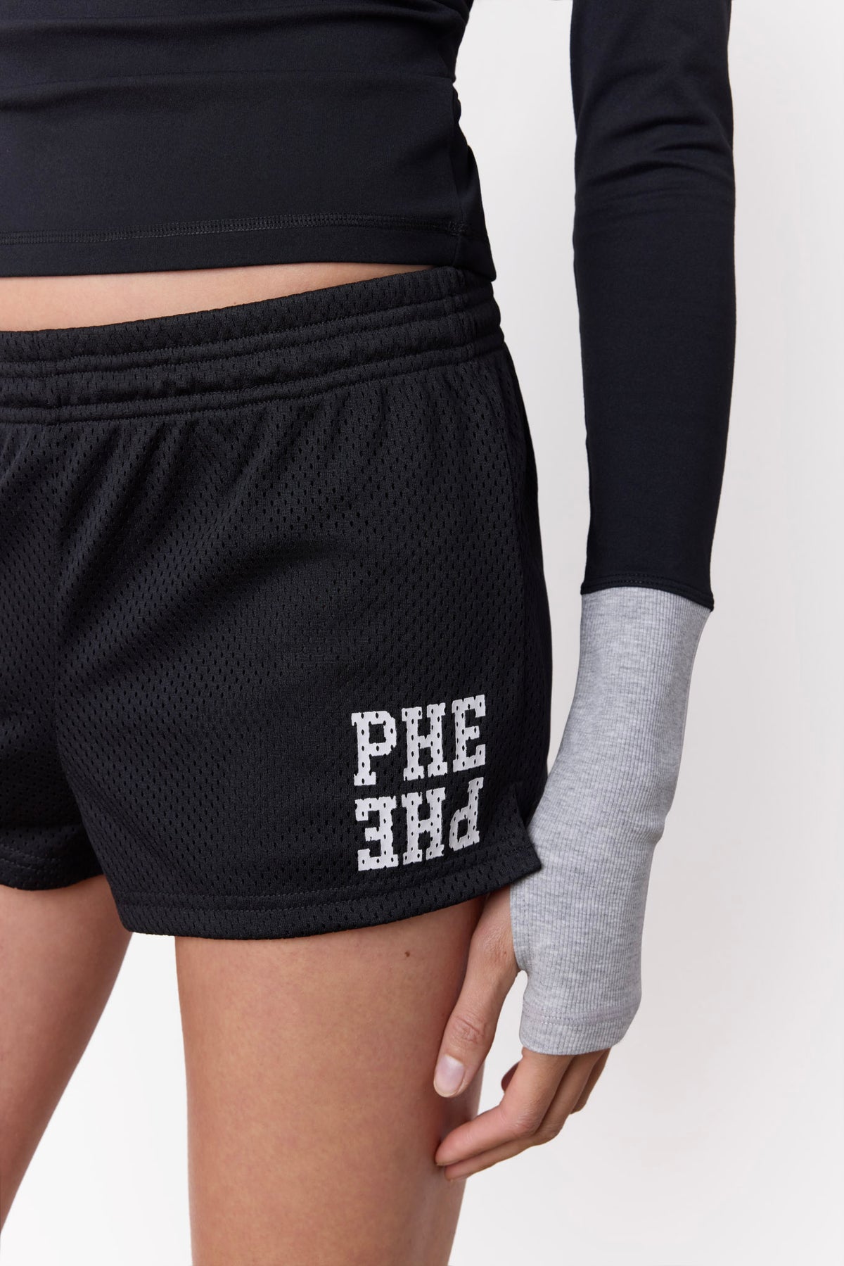 Black athletic mesh shorts with white 'PHE PHE' lettering on the lower left leg, shown on a model wearing a black long-sleeve top with grey cuffs and thumbhole sleeve against a plain white background.