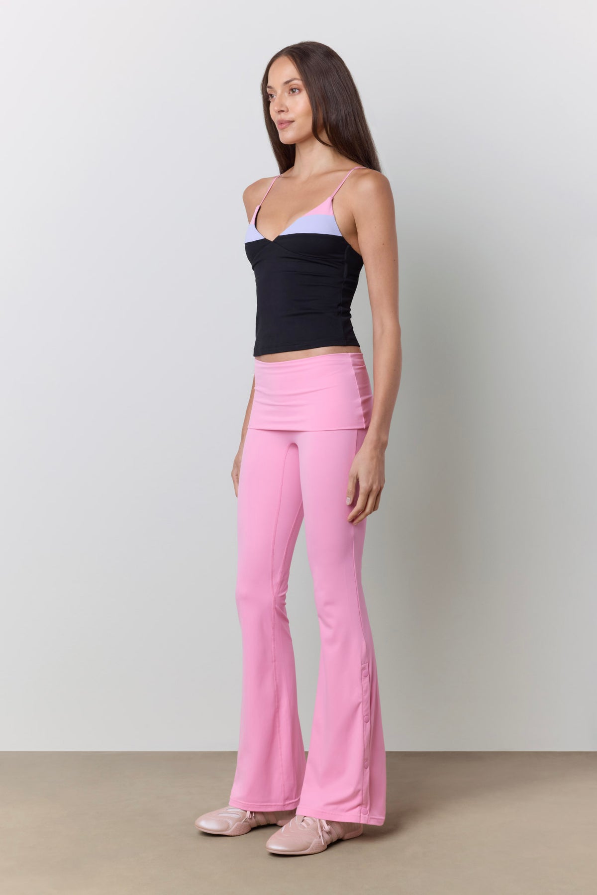 A woman modeling a pair of bright pink high-waisted flared pants, paired with a black camisole and light pink shoes, against a neutral studio background.