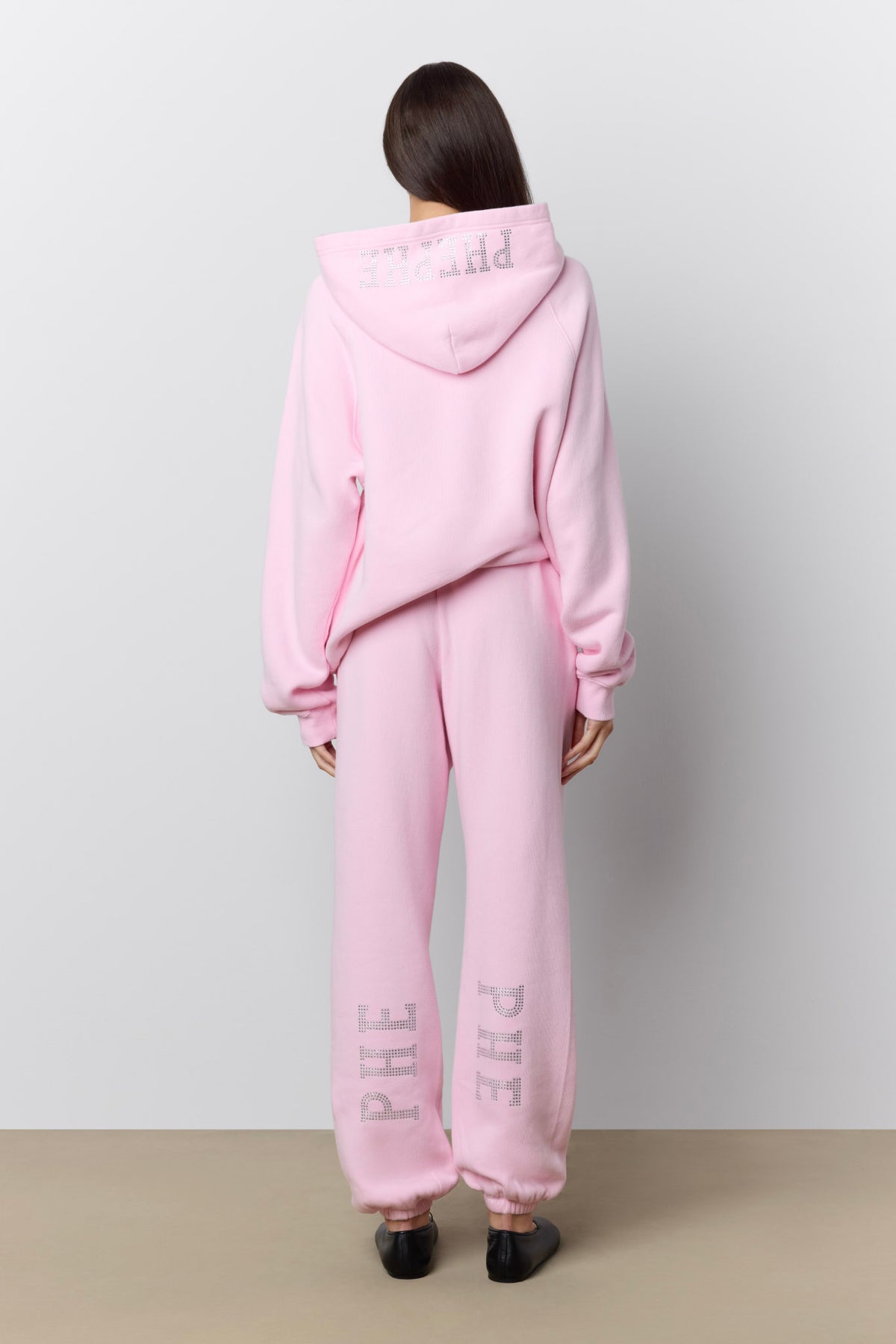 A model viewed from the back wearing a light pink matching lounge set consisting of a hooded sweatshirt and jogger pants, both featuring rhinestone 'PHE' lettering. The model is standing against a neutral light gray wall and beige floor.