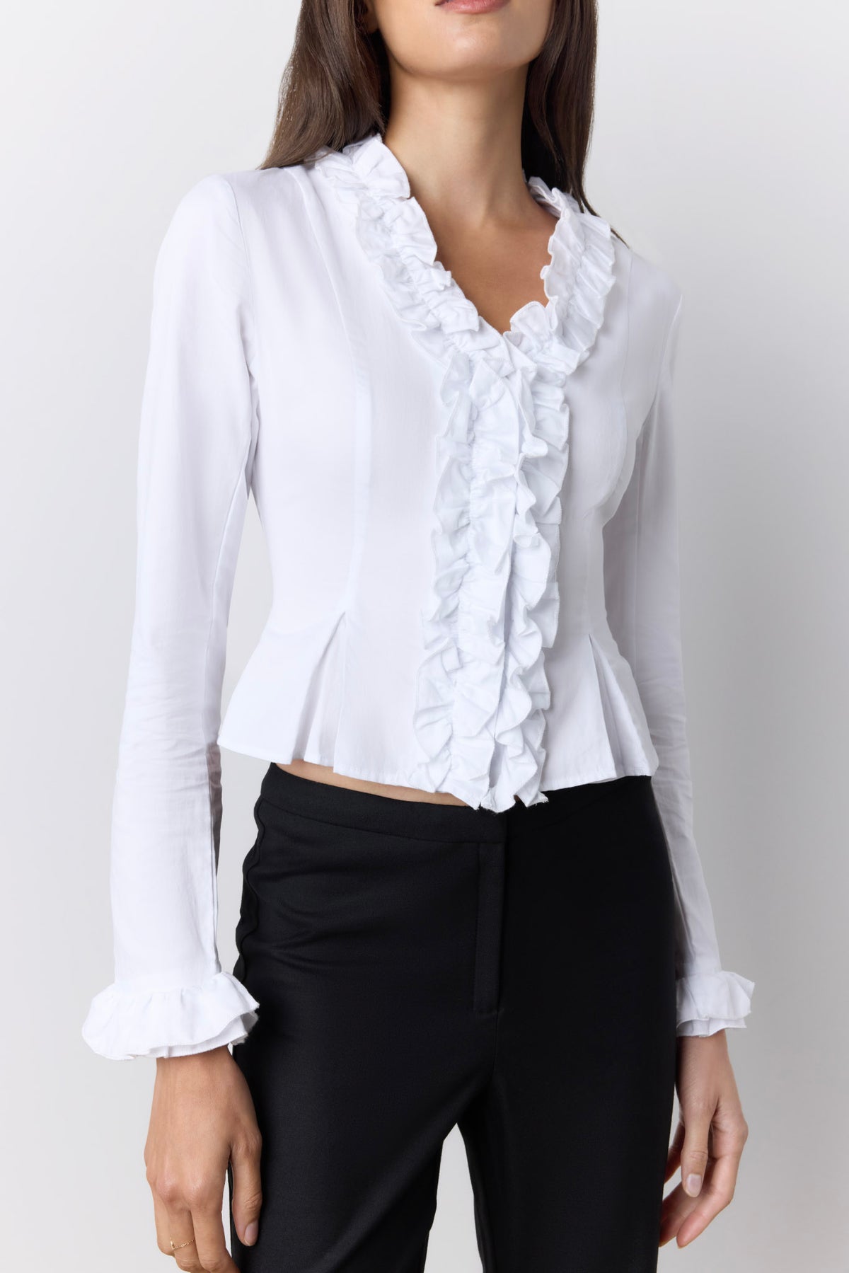 A woman wearing a white long-sleeve blouse with a ruffled V-neckline and ruffle trim down the front, paired with black trousers, against a light gray studio background.