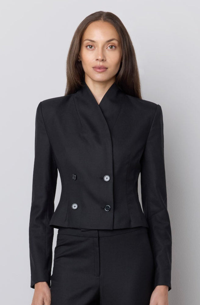 A model stands against a light gray studio wall wearing a tailored black two-piece suit with a structured double-breasted jacket and matching wide-leg trousers.