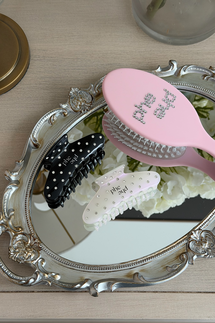 Two claw hair clips with rhinestones and the text "phe phe" are arranged on an ornate silver mirror, alongside a pink hairbrush with "phe phe" rhinestone lettering on a light wood tabletop.