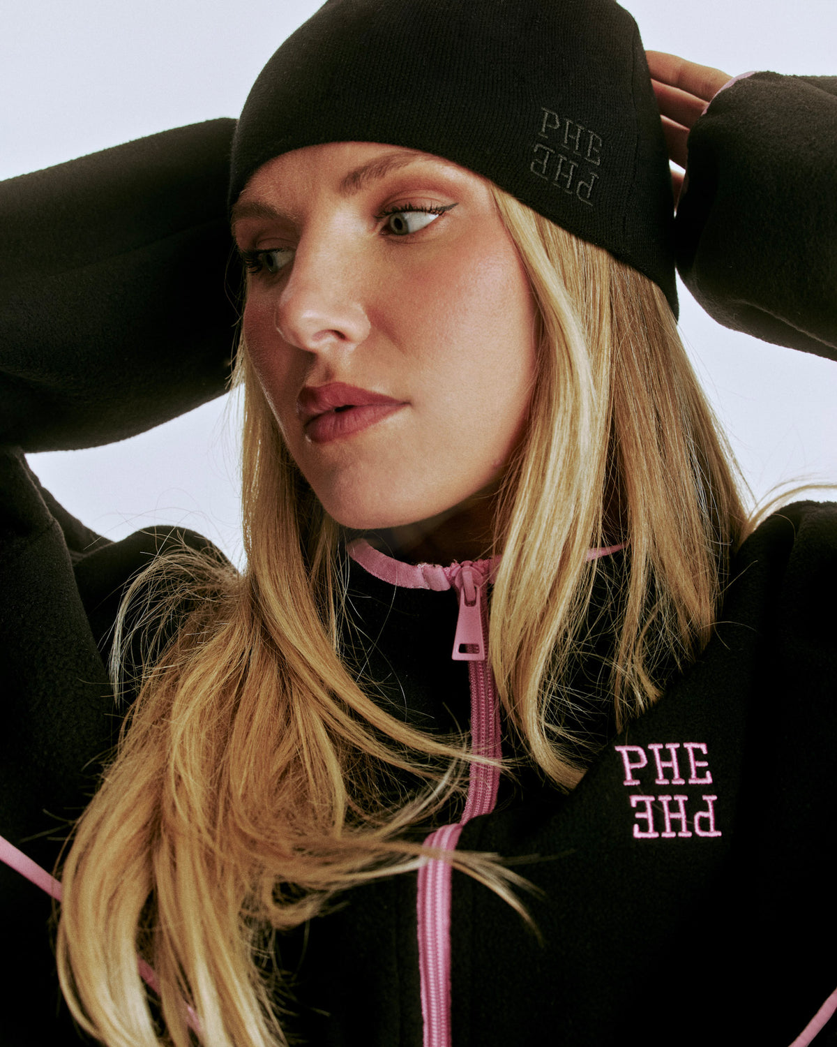 A blonde woman wearing a black beanie and black fleece jacket with pink zippers and pink embroidered 'PHE PHE' lettering, posed against a light gray background.