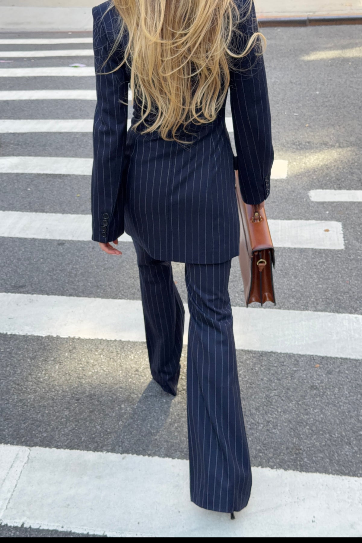 A woman viewed from behind walking across a crosswalk, wearing a navy blue pinstripe blazer dress and matching flared pants, and carrying a brown handbag.