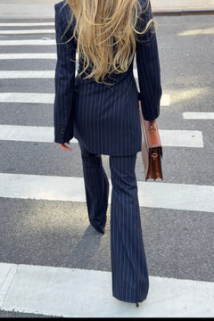A woman viewed from behind walking across a crosswalk, wearing a navy blue pinstripe blazer dress and matching flared pants, and carrying a brown handbag.
