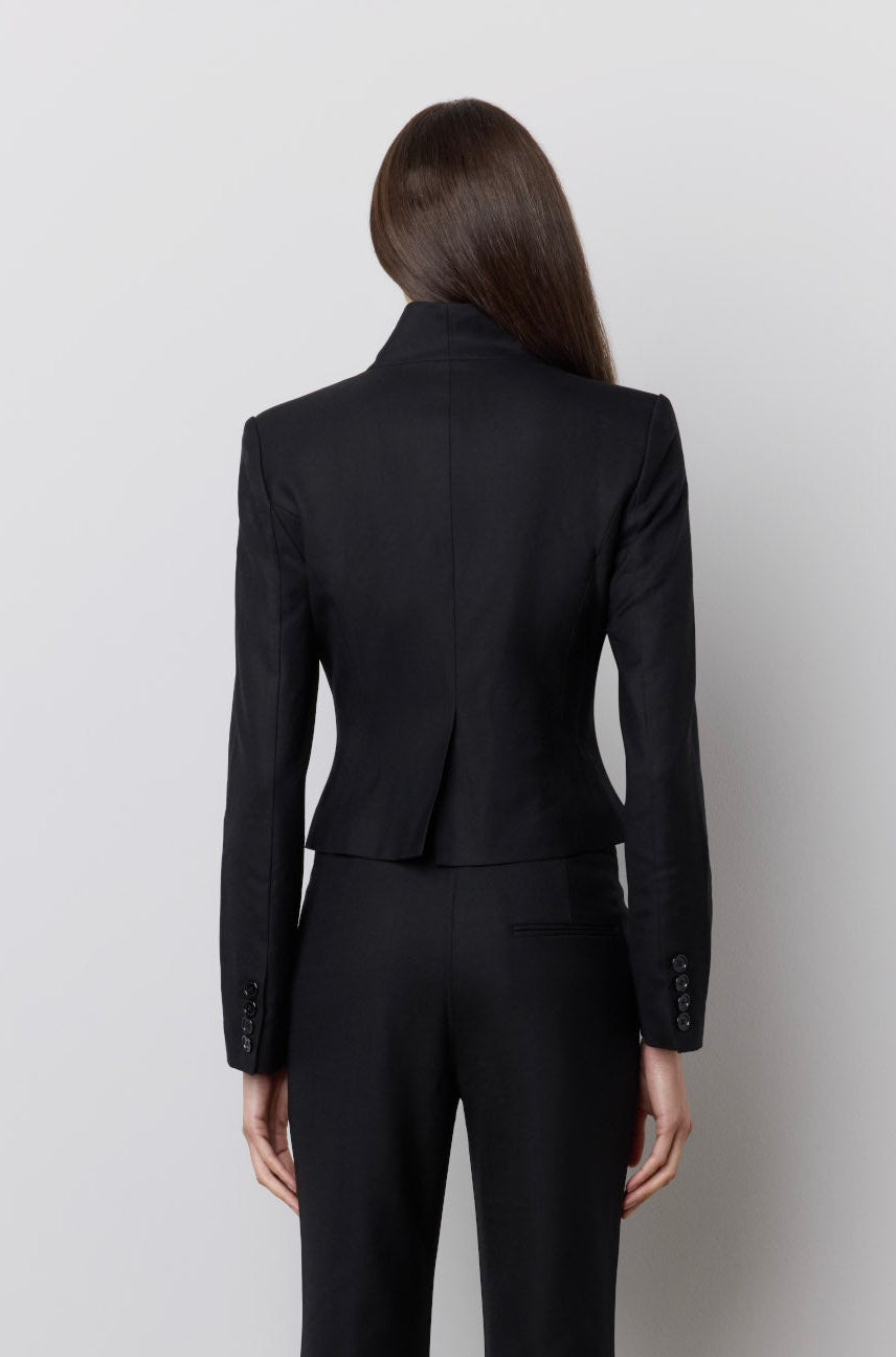 A model viewed from the back wearing a black tailored jumpsuit with long sleeves and flared pants, standing against a plain light gray wall.
