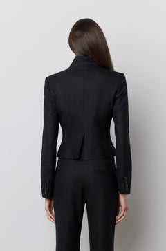A model viewed from the back wearing a black tailored jumpsuit with long sleeves and flared pants, standing against a plain light gray wall.