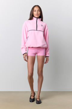 A model stands against a light gray studio background wearing a pink quarter-zip sweatshirt and matching pink shorts, with black shoes.