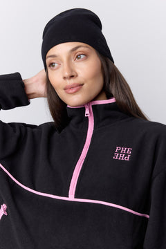 A woman wearing a black beanie and a black quarter-zip fleece jacket with pink zipper trim and pink embroidered lettering on the chest, photographed against a light gray studio background.