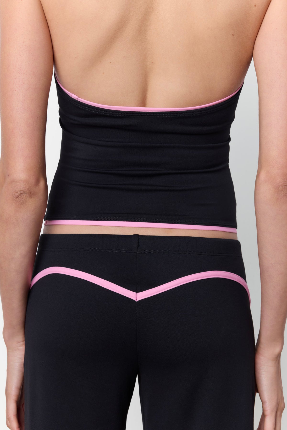 Rear view of a model wearing black athletic bottoms with a pink V-shaped trim and a matching black wrap-style top with pink trim, against a light gray studio background.