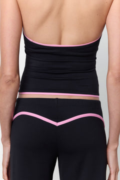 Rear view of a model wearing black athletic bottoms with a pink V-shaped trim and a matching black wrap-style top with pink trim, against a light gray studio background.