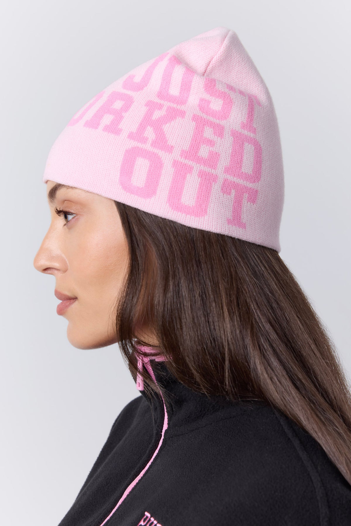 A woman in profile wearing a light pink knit beanie with the words 'JUST WORKED OUT' in darker pink lettering, paired with a black top with pink piping, against a light gray studio background.