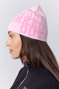 A woman in profile wearing a light pink knit beanie with the words 'JUST WORKED OUT' in darker pink lettering, paired with a black top with pink piping, against a light gray studio background.