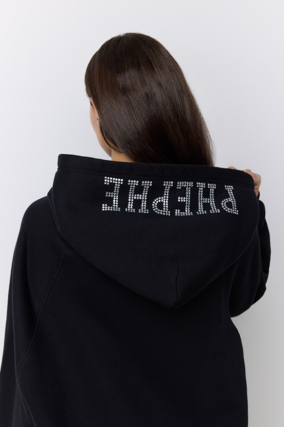 A person shown from behind wearing a black hoodie with the word 'PHEPHE' spelled out in sparkling silver rhinestones across the back of the hood, against a plain light gray background.