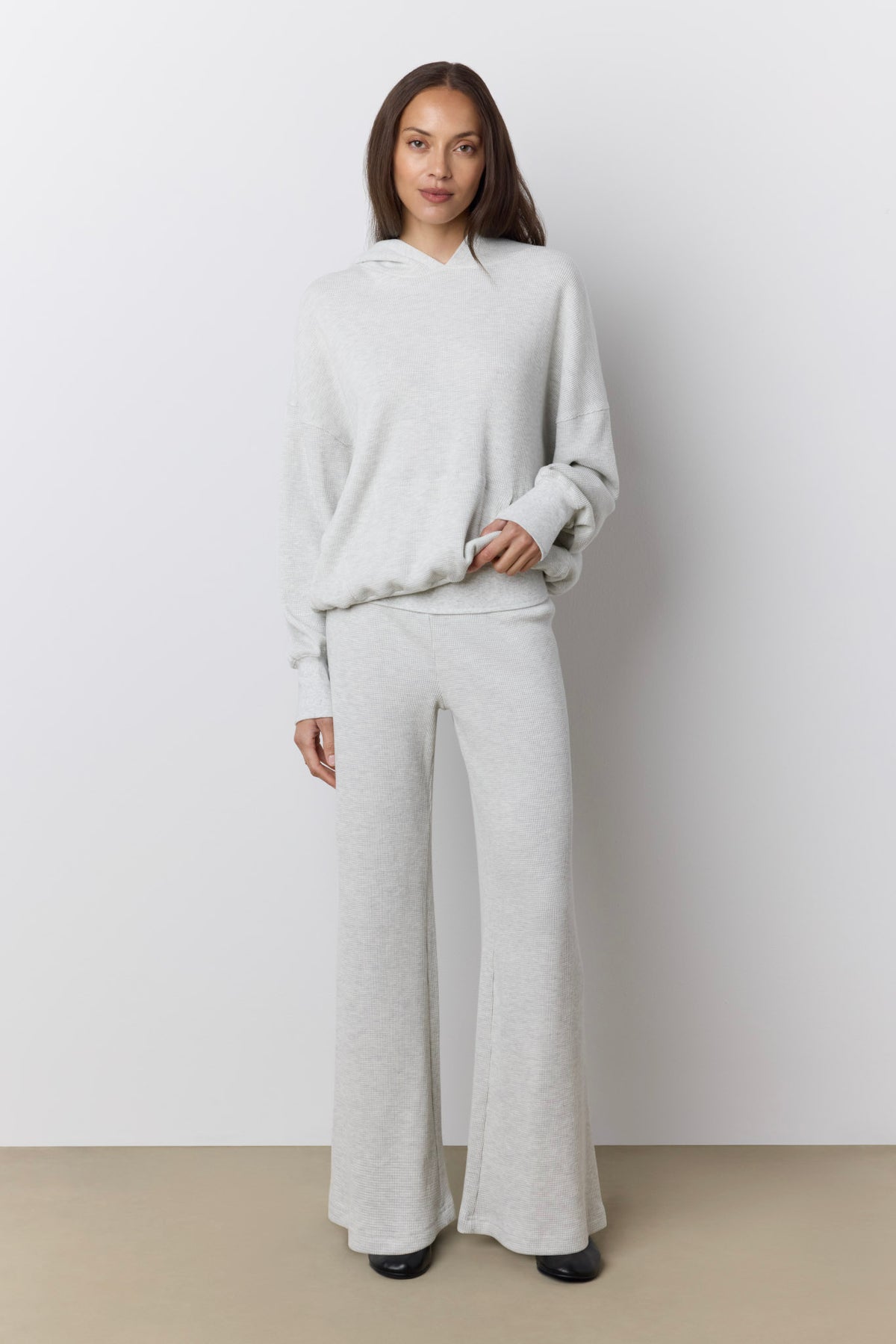 A woman modeling a light gray two-piece loungewear set consisting of a waffle knit long-sleeve hoodie and matching wide-leg pants, standing against a neutral studio background.