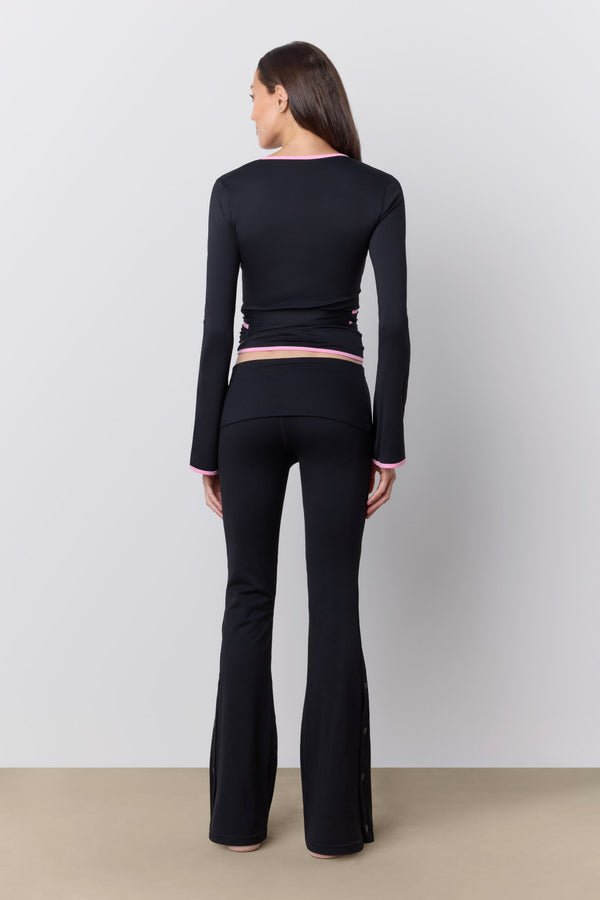 A woman shown from the back wearing a black long-sleeve wrap top with pink trim and matching black flared pants, standing against a light gray studio wall. |M1