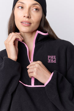 A woman wearing a black fleece quarter-zip pullover with pink trim and the letters 'PHE PHE' embroidered in pink on the chest, paired with a black beanie against a light gray background.