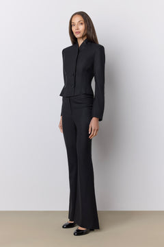 A model wearing a black tailored two-piece suit with a structured jacket and matching wide-leg trousers, standing against a light gray studio wall.