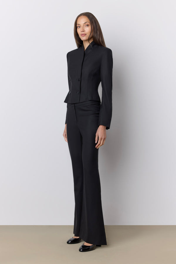 A model wearing a black tailored two-piece suit with a structured jacket and matching wide-leg trousers, standing against a light gray studio wall. |M1