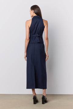 A woman viewed from the back wearing a sleeveless navy vest with thin vertical white pinstripes, a high neckline, and a matching fabric tie belt at the waist. She is standing against a plain light gray studio wall on a beige floor.