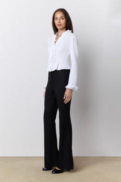 A woman modeling a white long-sleeve blouse with ruffled trim and black wide-leg trousers against a plain light gray studio background.
