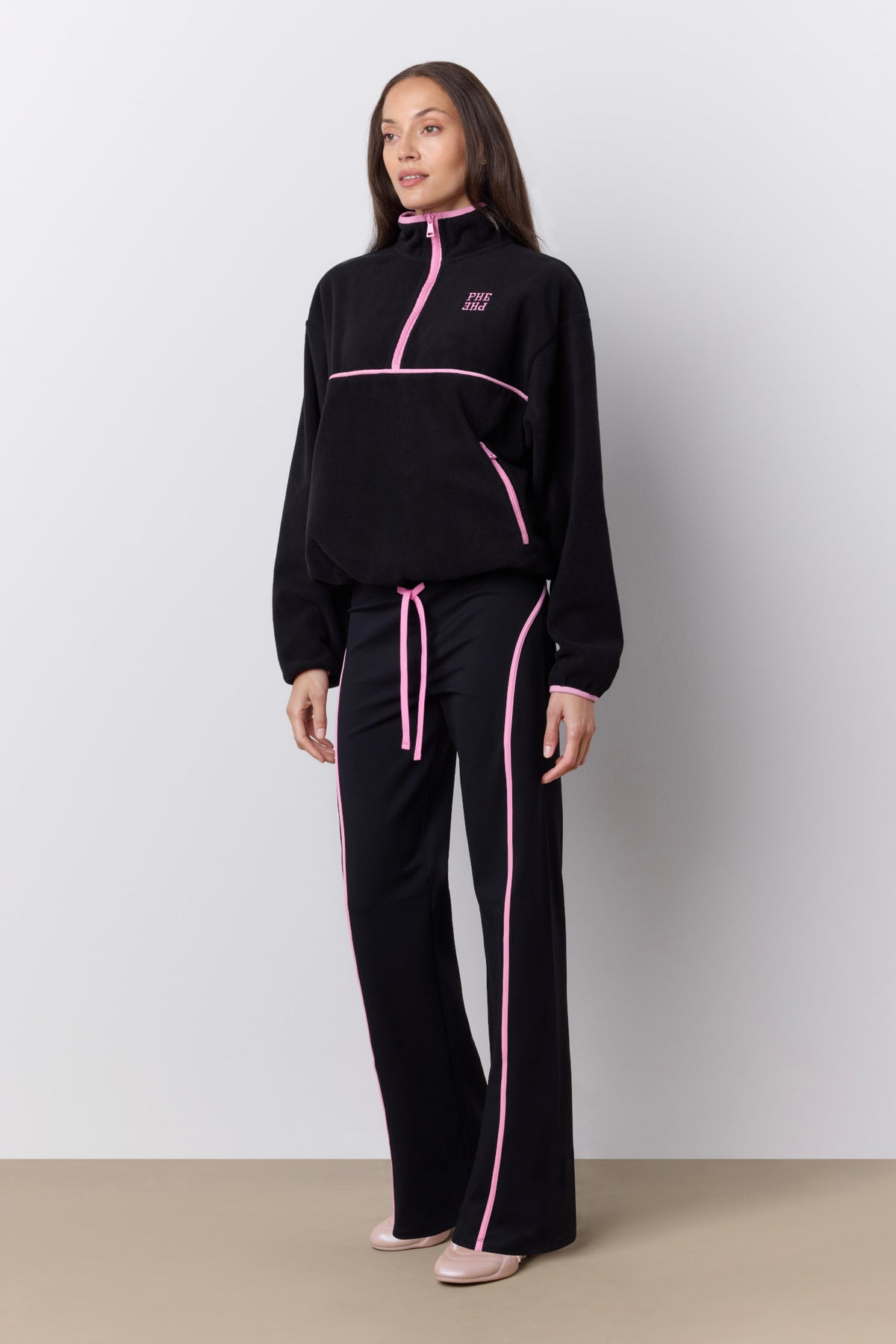 A woman modeling a black two-piece tracksuit with bright pink accents, including a half-zip jacket and matching wide-leg pants with pink piping and a drawstring, set against a neutral studio background.