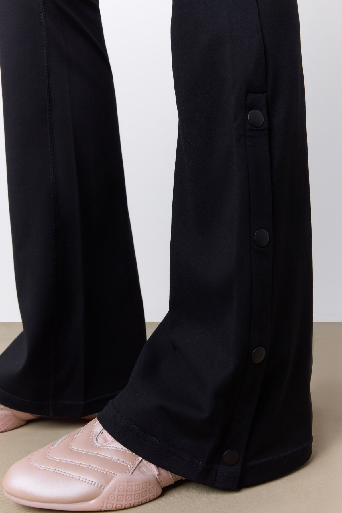 Close-up of black wide-leg trousers with dark buttons down the outer side, shown on a model wearing pale pink slip-on shoes against a neutral studio background.