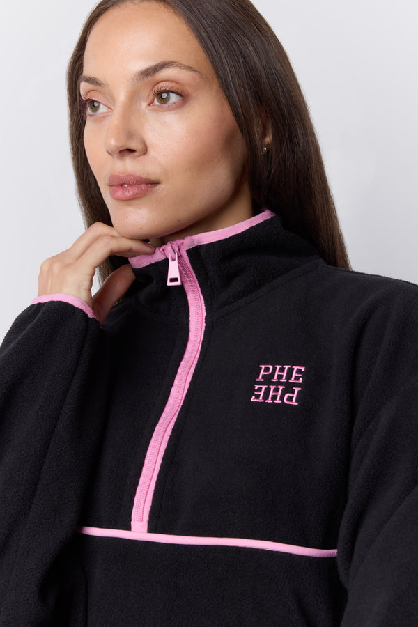 A woman wearing a black fleece half-zip jacket with pink trim and pink embroidered 'PHE' lettering on the chest, photographed against a light gray studio background. |M1