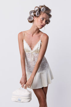 A woman wearing a white lace chemise and phe phe Hair Roller Set Pearl in her hair holds a small vanity case.