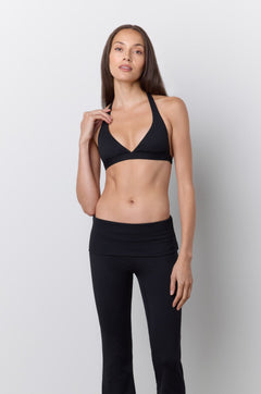 A woman modeling a black two-piece activewear set consisting of a halter-neck sports bra and matching high-waisted flared leggings, standing against a light gray studio wall.