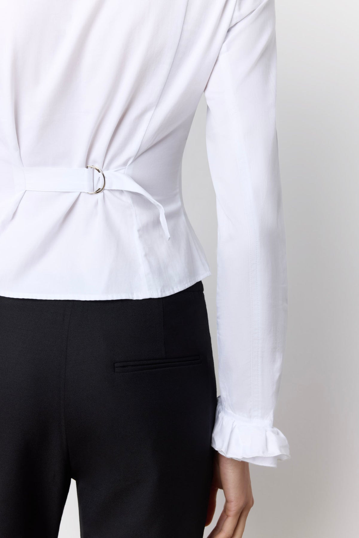 A close-up of a model wearing a white long-sleeve blouse with a tie waist detail and ruffled cuffs, paired with black trousers, shown from the back against a light gray background.