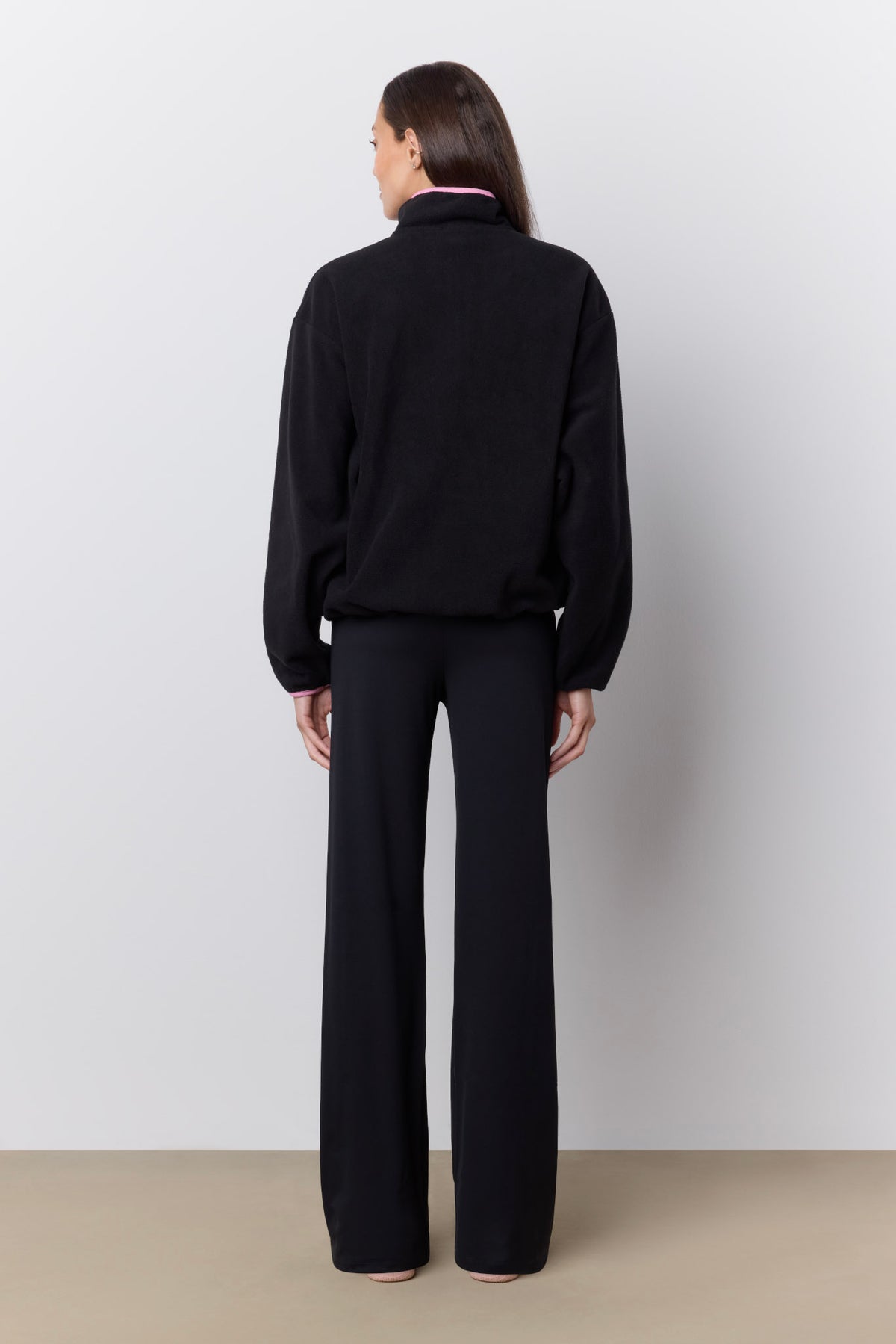 A model viewed from the back wearing a black long-sleeve mock-neck top and matching black wide-leg trousers, standing against a light gray wall and beige floor.