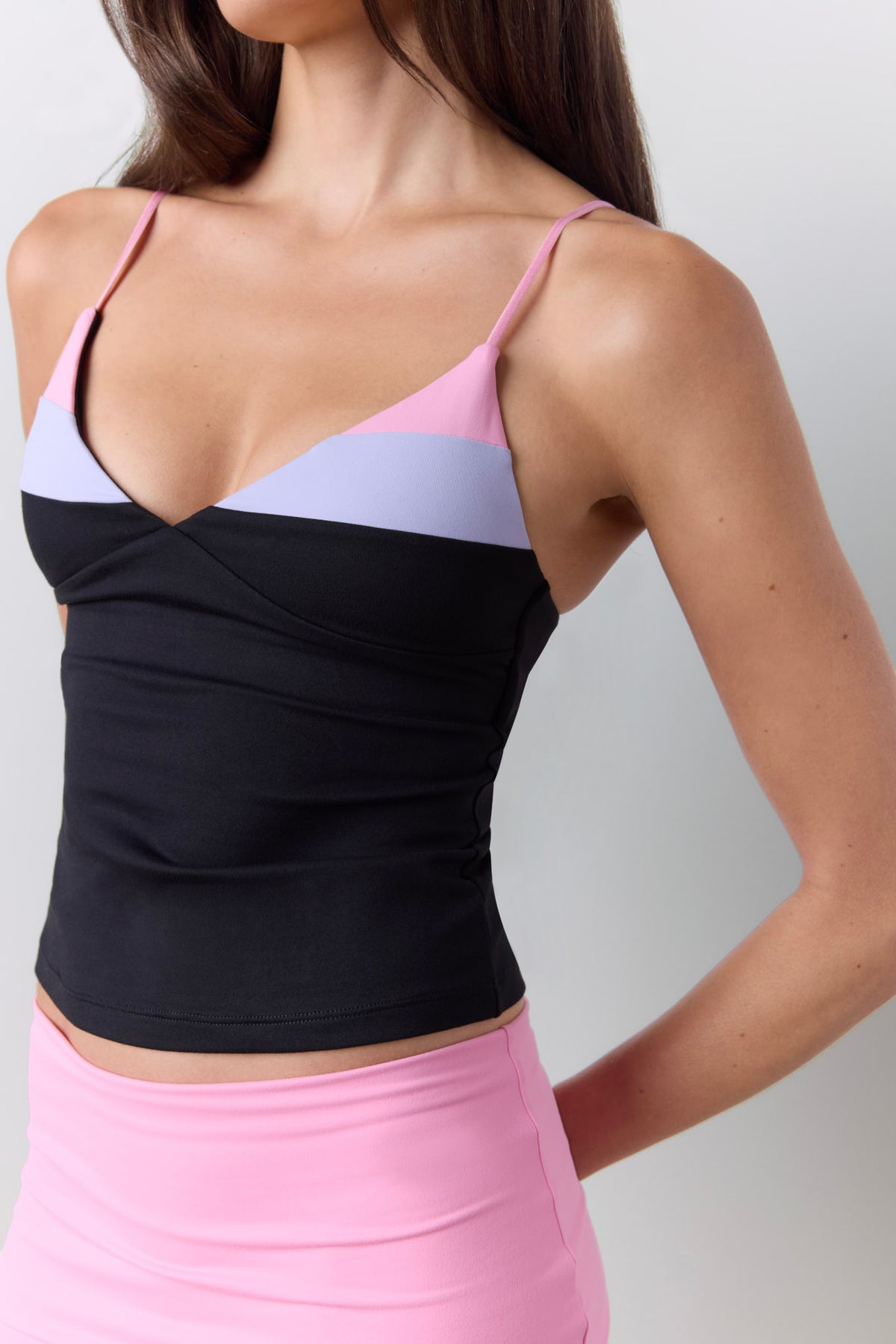 A close-up of a woman wearing a color-blocked camisole top with thin straps, featuring black, light purple, and pink sections, paired with a pink shorts against a light gray background.