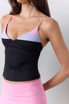 A close-up of a woman wearing a color-blocked camisole top with thin straps, featuring black, light purple, and pink sections, paired with a pink shorts against a light gray background.