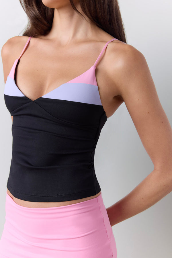 A close-up of a woman wearing a color-blocked camisole top with thin straps, featuring black, light purple, and pink sections, paired with a pink shorts against a light gray background. |M1