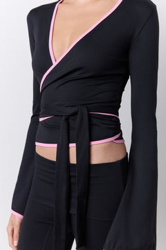 A close-up of a model wearing a black long-sleeve wrap-style top with pink trim and side cutouts, paired with black bottoms against a light gray studio background.