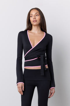 A woman modeling a black wrap-style long-sleeve top and matching flared pants set with pink trim details, standing against a light gray studio background.