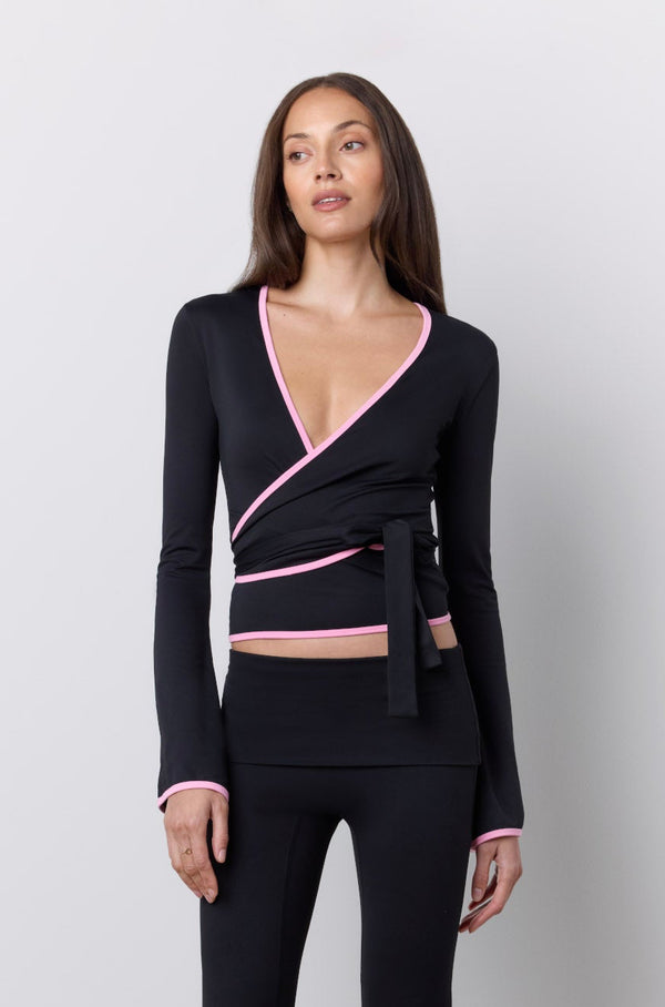 A woman modeling a black wrap-style long-sleeve top and matching flared pants set with pink trim details, standing against a light gray studio background. |M1