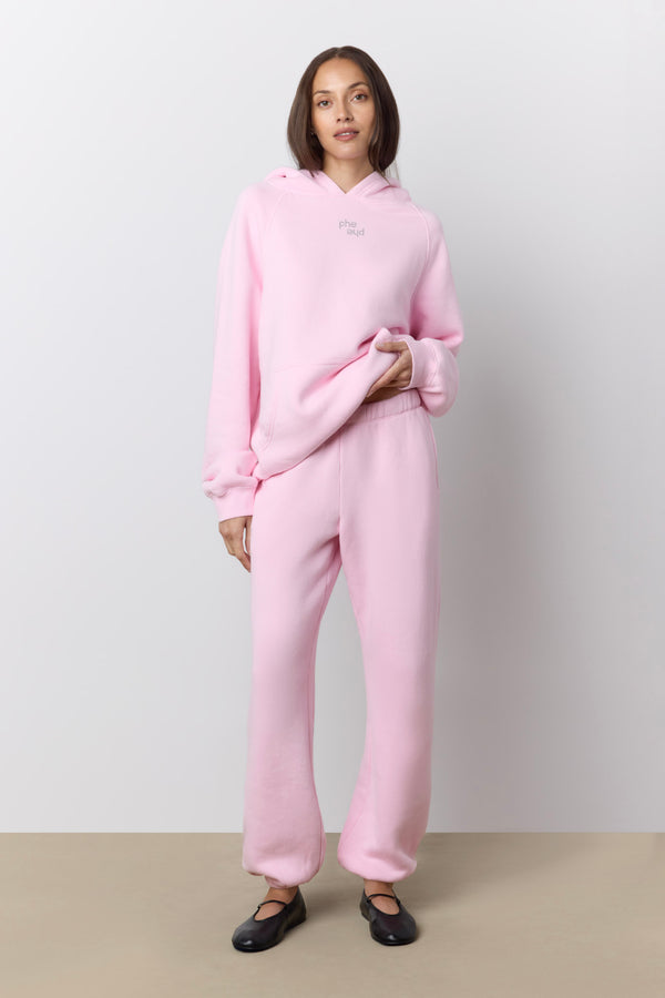 A woman modeling a light pink two-piece loungewear set consisting of a pullover hoodie and matching sweatpants, paired with black ballet flats, against a neutral studio background. |M1