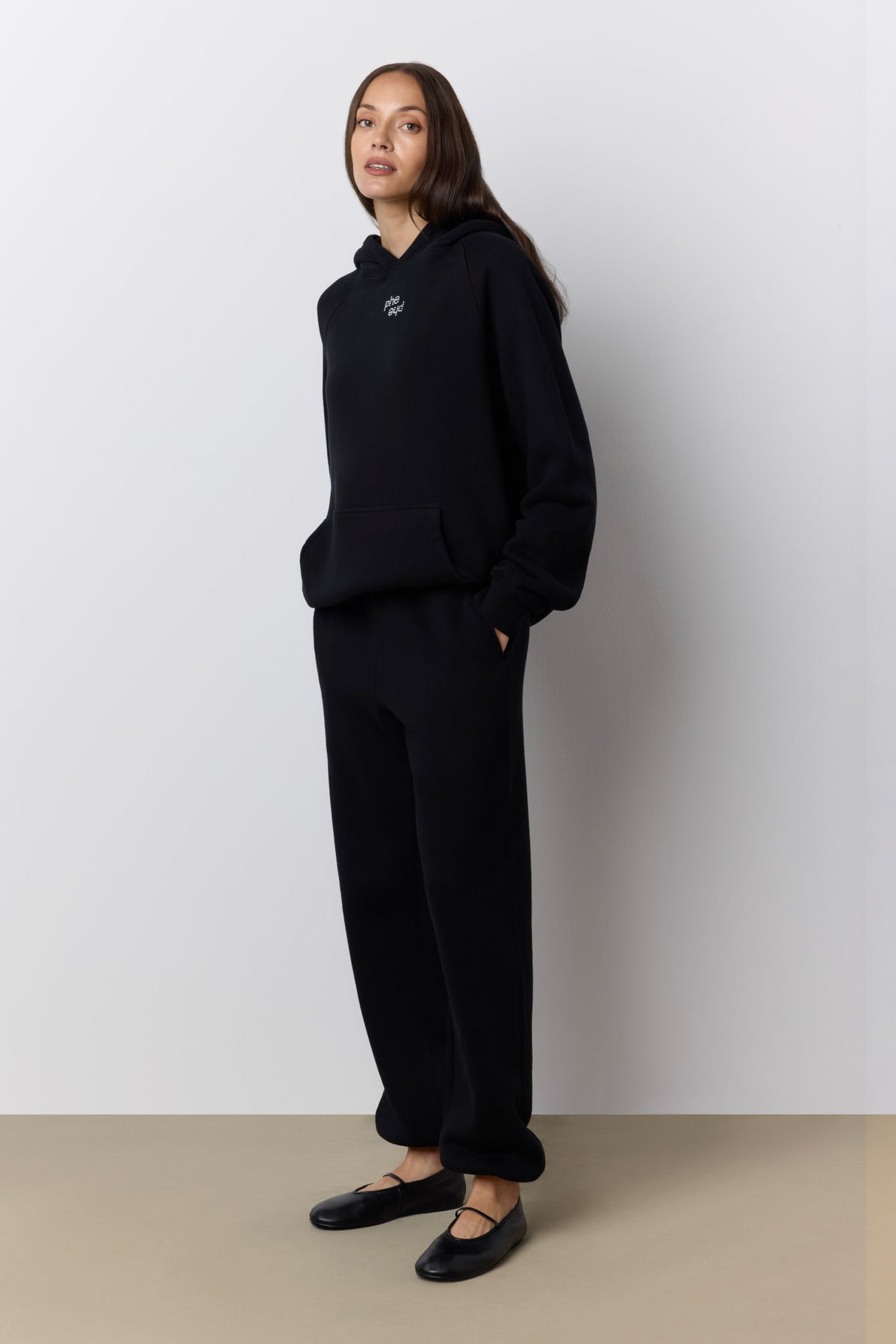 A woman modeling a black hooded sweatshirt and matching black wide-leg sweatpants against a light gray wall and beige floor.